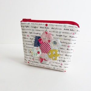 Puede incluir: Pequeño neceser de tela blanca con cremallera y ribete rojos. El neceser está decorado con un patchwork de piezas de tela hexagonales de colores. La tela está estampada con varias palabras y pequeñas ilustraciones.