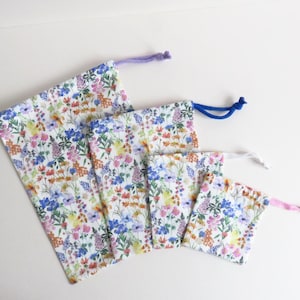 May include: Four drawstring bags in different sizes, arranged diagonally. The bags are white with a colourful floral pattern of blue, pink, yellow, and orange flowers. Each bag has a different coloured drawstring: purple, blue, white, and pink.