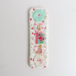 Puede incluir: Marcapáginas de tela hecho a mano sobre fondo blanco. Presenta hexágonos con motivos florales, cerezas y un oso de peluche. El texto "Fabric Textile Stitch Butt" es visible en el marcapáginas.