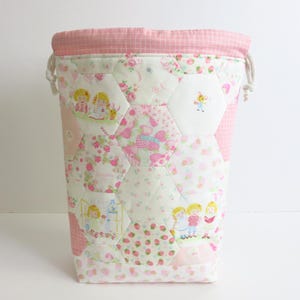 Puede incluir: Una cesta de almacenamiento de tela acolchada hecha a mano. La cesta presenta una combinación de colores rosa y blanco con un cierre de cordón. El exterior está decorado con parches hexagonales con patrones florales y de fresas, e ilustraciones de niños.