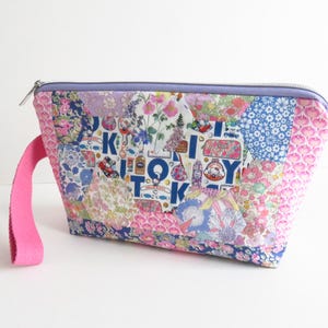 Op de afbeelding: Een patchwork make-up tasje met een roze handvat. De tas heeft een verscheidenheid aan bloemen- en patroonstoffen in roze, blauwe en witte tinten. De tas heeft een lavendelkleurige rits en het woord "K" is zichtbaar aan de voorkant.