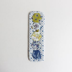 以下が含まれることがあります： 白の背景と青の幾何学模様の長方形の布製ブックマーク。緑、紫、青の色合いの、模様入りの六角形の布地が6つ、縦に並んでいます。