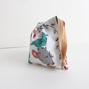 Pode incluir: Uma bolsa de cordão branco com um padrão floral com pássaros e folhas. A bolsa tem um cordão de cor castanho.