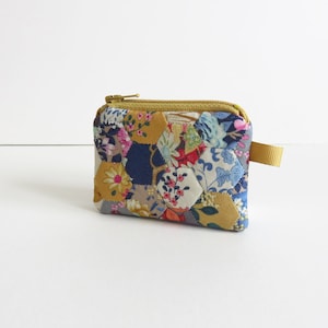 May include: A small, quilted zipper pouch with a floral pattern in shades of blue, pink, yellow, and green. The pouch has a gold zipper and a gold strap.