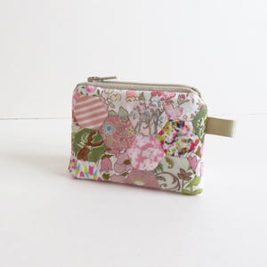 May include: A small, zippered pouch made of patchwork fabric. The fabric is a white background with pink, green, and brown floral patterns. The pouch has a beige zipper and a beige pull tab.