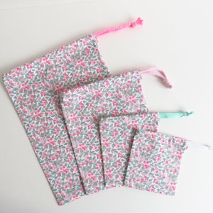 Può includere: Quattro sacchetti di stoffa con coulisse di diverse dimensioni, disposti in fila. Le borse presentano uno sfondo bianco con un motivo floreale rosa e verde. Ogni borsa ha un cordoncino di colore diverso: rosa, rosa chiaro, verde chiaro e bianco.