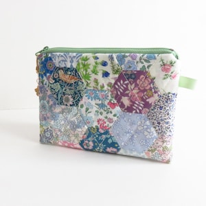 Puede incluir: Un neceser acolchado con un diseño de patchwork con estampados florales y de pájaros en varios colores. El neceser tiene una cremallera verde y un pequeño lazo verde en el lateral. El patchwork está hecho de formas hexagonales.