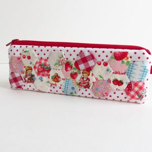 May include: A rectangular pencil case with a red zip and pull tab. The case is white with red polka dots and features a patchwork design of hexagonal fabric pieces with strawberry and cherry patterns. The patchwork includes images of a girl and the word "machine".