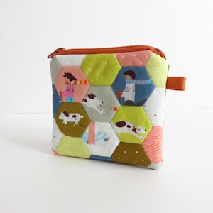 May include: A small, square fabric pouch featuring a patchwork design of hexagonal shapes. The fabric displays colorful illustrations of dogs and people in various colors including lime green, blue, brown, and white, with an orange zipper closure.