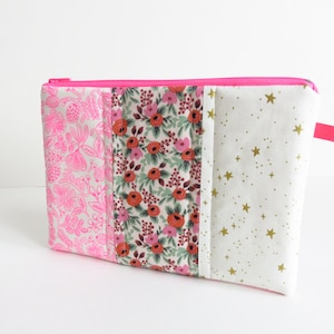 Może przedstawiać: Prostokątna kosmetyczka zapinana na zamek błyskawiczny z jasnoróżowym zamkiem i uchwytem. Kosmetyczka wykonana jest z trzech paneli materiału: różowego nadruku kwiatowego, kwiatowego wzoru z czerwonymi i różowymi kwiatami oraz białej tkaniny ze złotymi gwiazdami.