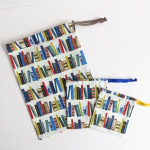 Puede incluir: Tres bolsas de cordón con fondo blanco y un patrón colorido de libros. La bolsa más grande tiene un cordón marrón, la bolsa mediana tiene un cordón azul y la bolsa más pequeña tiene un cordón amarillo.