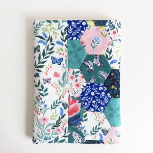 Pode incluir: Uma capa de caderno de tecido branco com um design de patchwork que apresenta formas hexagonais em tons de rosa, azul e verde. O tecido tem um padrão floral com borboletas.