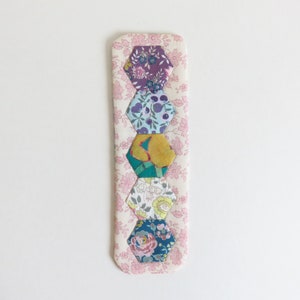 以下が含まれることがあります： ピンクの布製のしおりで、6つのカラフルな六角形の布のパッチが並んでいます。パッチは、紫、青、黄、緑、ピンクの色合いです。