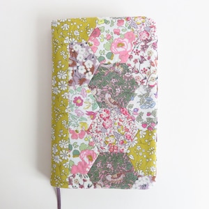 Può includere: Una copertina per quaderno in tessuto giallo con un design a patchwork di motivi floreali esagonali in tonalità di rosa, verde e bianco.