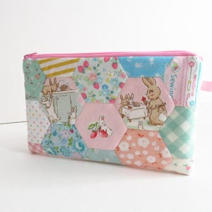 May include: A colorful, patchwork zippered pouch featuring hexagonal fabric panels. The panels display various patterns, including floral prints, polka dots, and illustrations of rabbits. The zipper is pink, and a small pink loop is attached to the side.