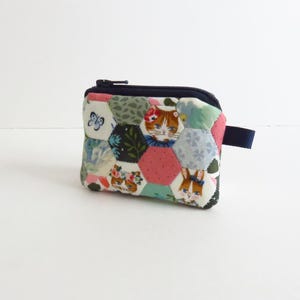 May include: A small, quilted coin purse with a navy blue zipper and pull tab. The patchwork design features hexagonal fabric pieces with floral, butterfly, cat, and rabbit patterns in pink, green, and blue hues.