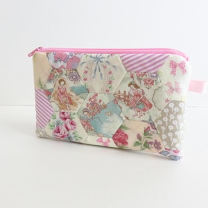 May include: A rectangular zippered pouch featuring a patchwork design of hexagonal fabric panels. The fabric displays various patterns including floral motifs, pink bows, diagonal stripes, and illustrations of figures in a garden setting.