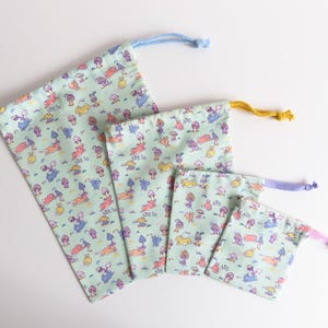 May include: Set of four drawstring bags in various sizes, all featuring a light green background with a repeating pattern of colorful mushrooms and animals. The bags are made of fabric and have a drawstring closure.
