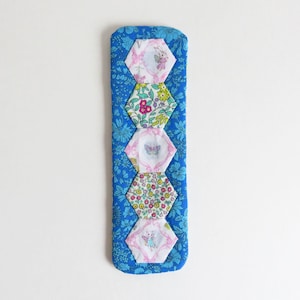 以下が含まれることがあります： 青い生地のブックマーク。花柄と6つの六角形のパッチが付いています。パッチには、ピンク、白、緑の蝶と花の模様があります。ブックマークの長さは約20cmです。