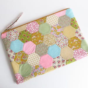 May include: A patchwork clutch bag featuring a pattern of hexagonal fabric pieces in various floral, striped, and dotted designs. The colour palette includes pink, blue, green, and yellow. It has a zip closure and a small wrist strap.