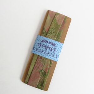 May include: Fabric strips in green and pink with a floral design, held together by a blue band. The band has the text "escapesq" and a website address. The fabric is presented on a brown cardboard backing.