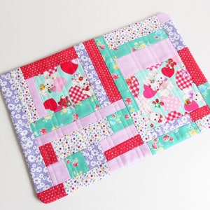 Puede incluir: Un mantel individual de patchwork con un patrón floral rosa, verde, rojo y blanco. El mantel individual presenta un patrón repetitivo de corazones y conejos.
