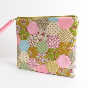 May include: A quilted pouch with a patchwork design. The pouch features a variety of hexagonal fabric pieces in pink, green, yellow, and blue with floral, polka dot, and striped patterns. It has a pink fabric pull tab and a gold zipper.