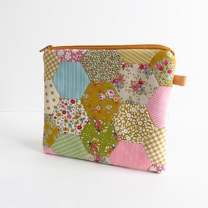 Könnte beinhalten: Ein Patchwork-Reißverschlusstasche mit verschiedenen floralen und geometrischen Mustern in Rosa, Gelb, Grün und Blau. Die Tasche hat einen braunen Reißverschluss und eine kleine Lederlasche. Das Design ist eine Mischung aus Vintage- und modernen Stilen.