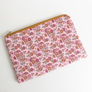 Puede incluir: Una bolsa rectangular rosa con cremallera y estampado floral. La bolsa presenta un patrón repetido de pequeñas flores en tonos rosa, rojo y naranja. La cremallera y el ribete son de color marrón dorado.