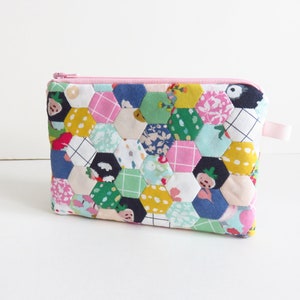 May include: A pink zippered pouch with a patchwork design made of colorful hexagonal fabric pieces. The fabric features various patterns including floral, geometric, and polka dots.