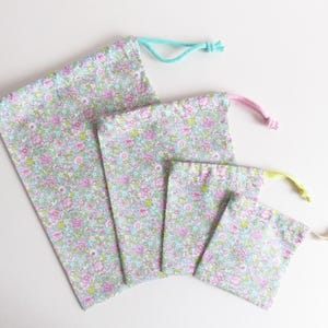 Pode incluir: Quatro sacos de cordão feitos de tecido com padrão floral em vários tamanhos. Os sacos têm cordões de diferentes cores: turquesa, rosa, amarelo e creme.