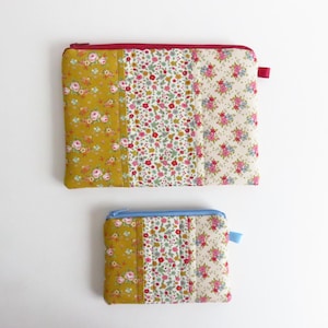 Könnte beinhalten: Zwei gesteppte Stoffbeutel. Der größere Beutel hat einen weinroten Reißverschluss und eine Lasche, und der kleinere Beutel hat einen blauen Reißverschluss und eine Lasche. Beide Beutel weisen florale Muster in Gelb, Rosa und Weiß auf.