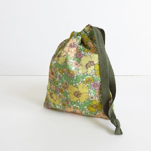 Op de afbeelding: Een klein trekkoordtasje van gele stof met een bloemenprint. De tas heeft een groen trekkoord.