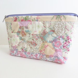 Op de afbeelding: Een tasje met rits voorzien van een patchworkontwerp van diverse bloemen- en patroonstoffen in tinten roze, crème en lichtblauw. De buitenkant is gemaakt van doorgestikte zeshoekige stoffen stukjes met een lavendelkleurige ritssluiting aan de bovenkant.