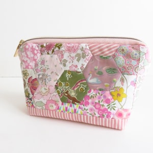 May include: Pink patchwork cosmetic bag with a zipper closure and a floral pattern. The bag is made of fabric and has a pink and white striped trim.