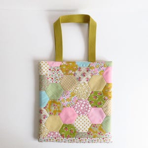 May include: A quilted tote bag with a patchwork design. The bag features a variety of hexagonal fabric pieces in pastel colors, including pink, yellow, green, and blue, with floral and dotted patterns. The handles are a solid olive green.