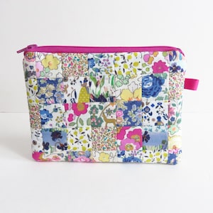 Puede incluir: Un neceser acolchado con diseño de patchwork. El neceser presenta una vibrante mezcla de estampados florales en rosa, azul, amarillo y verde. Tiene una cremallera rosa brillante y un pequeño lazo rosa.