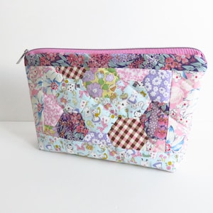 May include: A zipped pouch featuring a patchwork design of various floral, checkered, and character-themed fabric patterns in shades of pink, blue, purple, and brown. The top edge is finished with a pink and white striped fabric trim.