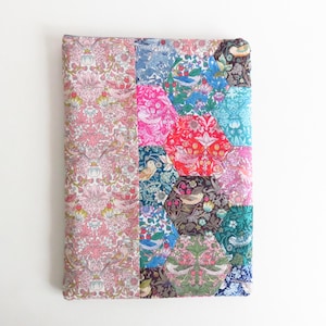 Puede incluir: Un cuaderno o diario cubierto de tela con un diseño de patchwork. El lado izquierdo presenta un patrón floral rosa, mientras que el lado derecho es una mezcla de parches hexagonales coloridos con motivos florales y de pájaros. La cubierta es rectangular.