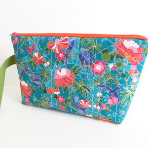 Op de afbeelding: Een gewatteerd tasje met rits op een blauwgroene achtergrond met een kleurrijk patroon van vogels, bloemen en bamboebladeren. Het tasje heeft een oranje ritssluiting aan de bovenkant en een lichtgroene stoffen polsband aan de zijkant.
