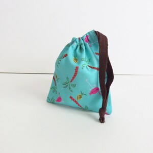 Peut inclure: Un sac à cordon turquoise avec un cordon marron. Le sac a un imprimé tropical avec des flamants roses, des palmiers et des ananas.