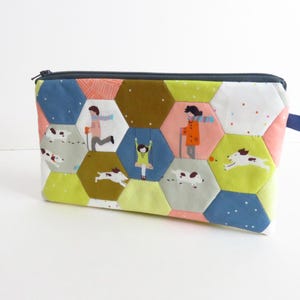 May include: A rectangular zippered pouch featuring a patchwork design of multicolored hexagons. The fabric pattern displays illustrations of people walking, a person on a swing, and dogs running against backgrounds of blue, green, and pink.