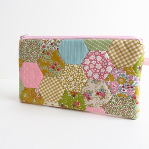 Może przedstawiać: Ręcznie robiona, pikowana kosmetyczka z różowym zamkiem błyskawicznym i uchwytem. Kosmetyczka wykonana jest z różnych tkanin w kwiaty i wzory w odcieniach różu, zieleni, żółci i niebieskiego, ułożonych w sześciokątny wzór. Kosmetyczka ma kształt prostokątny.