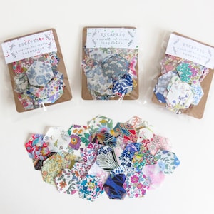 Op de afbeelding: Zeshoekige stofstukken in diverse bloempatronen. Drie pakketten met stofstukken worden weergegeven met de tekst "ESCAPESQ a passion for crafts handmade". Er wordt ook een stapel losse stofstukken getoond.