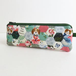 May include: A quilted pencil case with a green zipper and pull tab. The case is made of hexagonal fabric patches in various floral and animal patterns, including a dog, cats, and text that reads "Rose Viola Anemone."