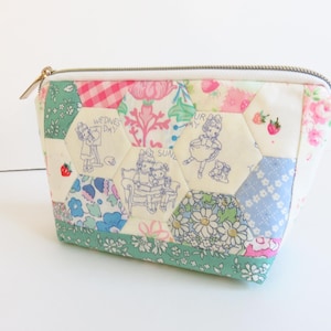 May include: A white zippered pouch with a patchwork design featuring hexagonal pieces of fabric in various floral patterns, including pink, blue, and green. The fabric pieces are sewn together to create a whimsical and colorful design. The pouch is perfect for storing small items like makeup, jewelry, or craft supplies.
