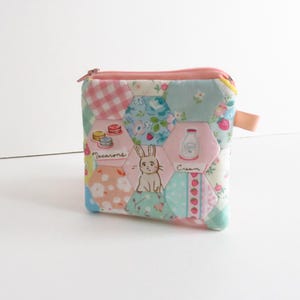 May include: A small, quilted coin purse with a pink zipper and pull tab. The patchwork design features hexagonal patterns in pastel colors, including pink, blue, and green. Illustrations of macarons, a bunny, and a milk bottle are included.