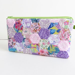 Puede incluir: Una bolsa de cremallera de patchwork morado y rosa con una cremallera verde y una lengüeta verde. La bolsa está hecha de tela con un patrón hexagonal.
