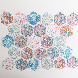 May include: A collection of twenty-five fabric hexagons arranged in a honeycomb pattern. Each hexagon features a distinct floral print in various colours including blue, pink, orange, and white, showcasing intricate botanical and geometric patterns.