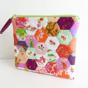 May include: A vibrant, quilted pouch with a green zip and a purple fabric pull. The pouch is constructed from hexagonal fabric pieces featuring diverse patterns, including floral, animal, and geometric designs. The pouch is approximately 20 cm wide and 15 cm tall.